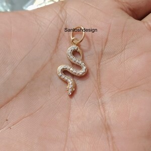 Puede incluir: Un colgante de serpiente en tono dorado con un cuerpo curvo en forma de S incrustado con pequeñas piedras claras y brillantes. El colgante incluye una anilla circular en la parte superior para su fijación y muestra el texto Santoshdesign.