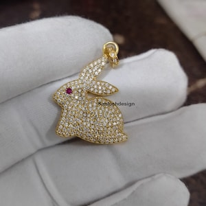May include: A gold-toned pendant in the shape of a rabbit, encrusted with small, clear stones. The rabbit's eye is a single red stone.