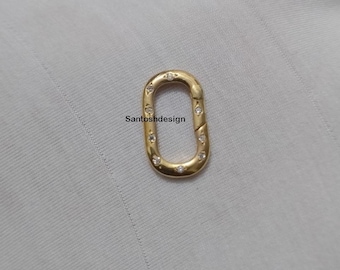 Puede incluir: Un cierre ovalado de color dorado con pequeños diamantes de imitación transparentes dispuestos alrededor del perímetro. El cierre está diseñado para conectar dos piezas de joyería.