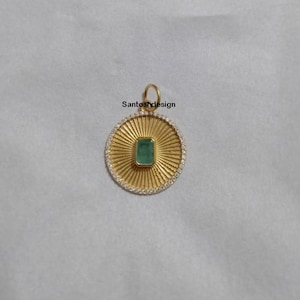 May include: A gold pendant with a green emerald gemstone set in a circular frame with a sunburst design and a border of small clear stones. The pendant is on a white background.