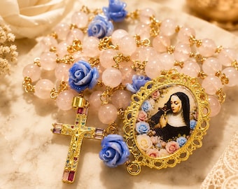 May include: A rosary featuring pale pink round beads, blue rose-shaped beads, and a gold-toned crucifix inlaid with multicolored rectangular stones. An oval gold-toned pendant displays a portrait of a nun holding a cross amidst floral accents.