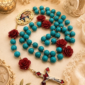 Peut inclure: Un chapelet avec des perles turquoise, des accents de roses rouges et un crucifix doré. Le chapelet comprend une image encadrée d'or et est posé sur un fond doux et neutre avec une bougie et une figurine d'ange.