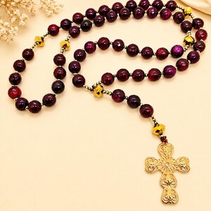 Puede incluir: Un rosario con cuentas esf&eacute;ricas de color p&uacute;rpura intenso intercaladas con cuentas m&aacute;s peque&ntilde;as facetadas en tono dorado met&aacute;lico. El collar termina con un colgante de cruz dorada de estilo filigrana con un dise&ntilde;o de metal calado detallado.