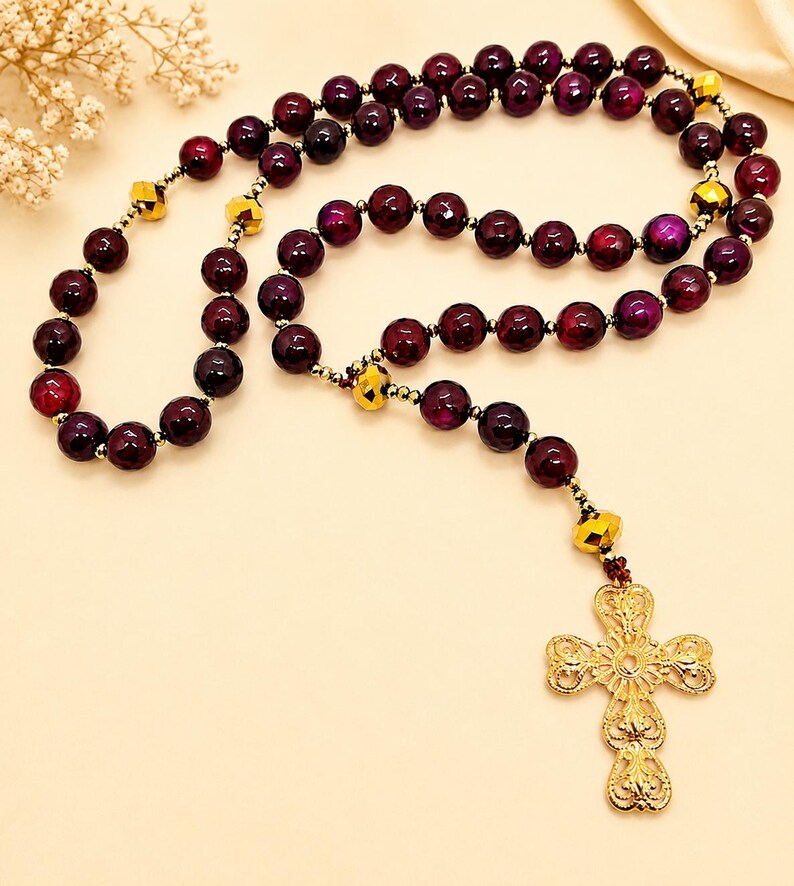 Puede incluir: Un rosario con cuentas esf&eacute;ricas de color p&uacute;rpura intenso intercaladas con cuentas m&aacute;s peque&ntilde;as facetadas en tono dorado met&aacute;lico. El collar termina con un colgante de cruz dorada de estilo filigrana con un dise&ntilde;o de metal calado detallado.