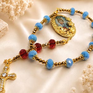 Könnte beinhalten: Ein goldfarbener Rosenkranz mit blauen und roten Perlen. Der Rosenkranz hat ein goldenes Kreuz mit klaren Kristallen und einen goldenen Anhänger mit einem religiösen Bild. Die Perlen sind facettiert und durch goldfarbene Glieder verbunden.