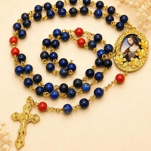 Può includere: Un rosario con grani in lapislazzuli blu scuro e perline rosse vivaci collegate da una catena in metallo dorato. Il pezzo include un crocifisso dorato e un elemento centrale ovale che raffigura un ritratto circondato da dettagli floreali.