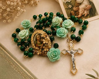 Può includere: Un rosario con perline verdi e accenti di rose verde chiaro. Il rosario presenta un crocifisso color oro e un medaglione centrale con un'immagine. Un libro vintage e una carta religiosa sono anche nell'immagine.