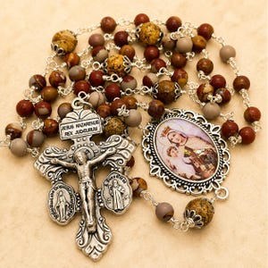 May include: A rosary featuring multi-colored brown and tan stone beads linked by silver-toned metal wire. The centerpiece includes a silver crucifix with the text JESUS NAZARENUS REX JUDAEORUM and an oval pendant depicting a figure with a child.