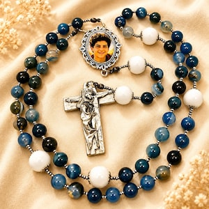 May include: A rosary featuring blue and black agate beads interspersed with larger white beads. The centrepiece is an oval metal frame containing a portrait of a young person, and the crucifix is a silver-toned metal cross with a detailed figure.