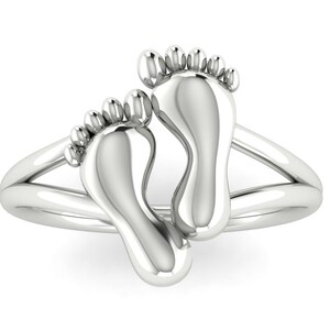 May include: A silver ring featuring two baby footprints. The ring's band splits into two strands that meet at the top, supporting the footprints. The footprints are detailed, showing toes and the shape of the feet. The ring is a symbol of new life.