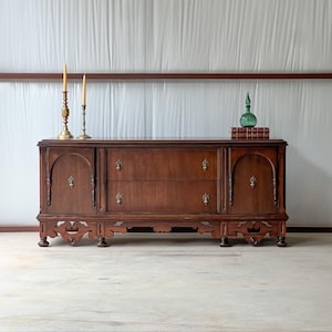 May include: A dark wood antique buffet with two drawers and two cabinet doors. The buffet has ornate brass hardware and decorative carvings. Two brass candlesticks with lit candles and a green glass bottle sit on top.