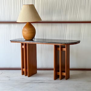 May include: A console table featuring a dark gray marble top and two vertical wooden support bases with a slatted design. A table lamp with a spherical, light brown textured base and a conical beige fabric shade sits on the table surface.