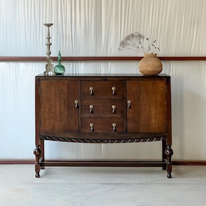 May include: A dark wood sideboard with three drawers and two cabinet doors. The sideboard has brass hardware and is decorated with a candle holder, a green glass bottle, and a woven vase with dried flowers.