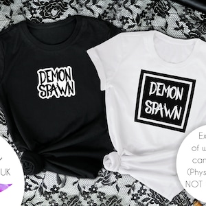 Puede incluir: Dos camisetas con el texto "DEMON SPAWN" en un diseño gráfico en blanco y negro. Las camisetas se muestran sobre un fondo de encaje negro.