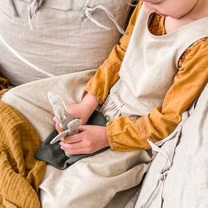 May include: A young person sits on a couch with a plush toy in their hands. The couch is covered in a light beige fabric and the person is wearing a light beige dress with a mustard yellow long-sleeved shirt underneath. The person is holding a gray plush toy with a brown nose and ears.