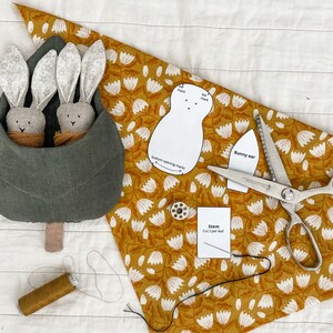May include: A green fabric bunny nest with two gray bunny plushies inside. The nest is on a white surface with a yellow floral fabric, a pair of pinking shears, a spool of thread, and a paper pattern for a bunny.