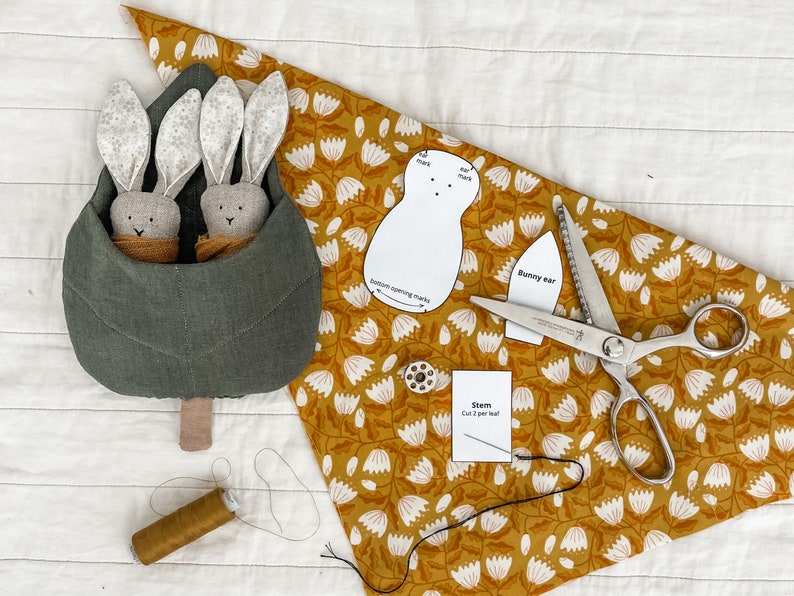 May include: A green fabric bunny nest with two gray bunny plushies inside. The nest is on a white surface with a yellow floral fabric, a pair of pinking shears, a spool of thread, and a paper pattern for a bunny.