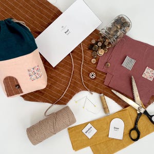 May include: A craft kit for making a house-shaped pouch. The kit includes fabric pieces in pink, green, brown, and white, along with buttons, twine, and a pattern sheet. The fabric pieces are labeled "Roof", "Door", and "Window".