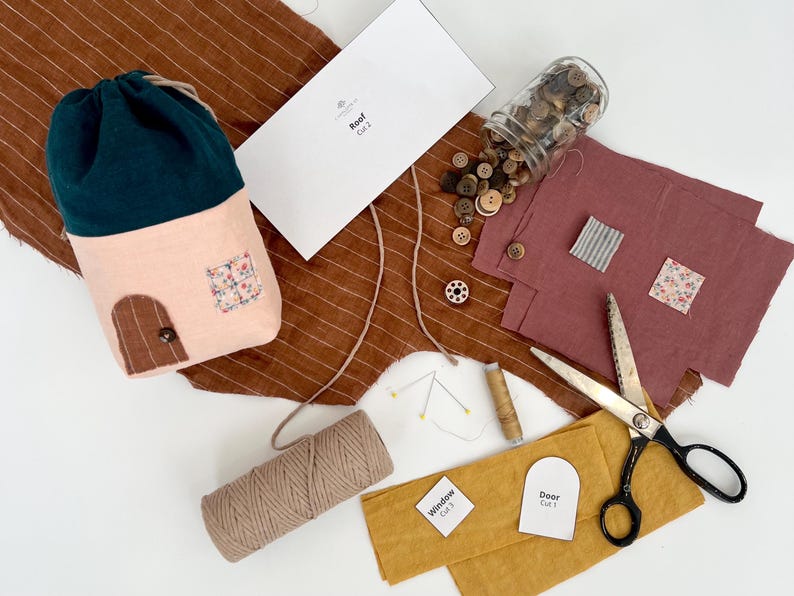 May include: A craft kit for making a house-shaped pouch. The kit includes fabric pieces in pink, green, brown, and white, along with buttons, twine, and a pattern sheet. The fabric pieces are labeled "Roof", "Door", and "Window".