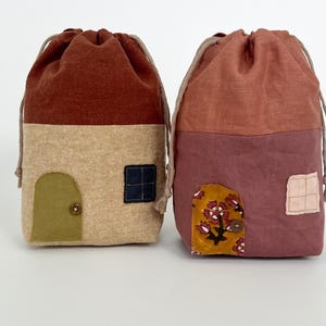 May include: Two fabric drawstring bags shaped like houses. The bag on the left is brown, beige, and green with a small door and window. The bag on the right is pink, beige, and yellow with a floral design and a window.