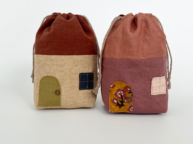 May include: Two fabric drawstring bags shaped like houses. The bag on the left is brown, beige, and green with a small door and window. The bag on the right is pink, beige, and yellow with a floral design and a window.