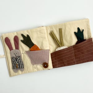 Puede incluir: Un libro de actividades de tela con un tema de conejos y jardín. El conejo lleva un chaleco a cuadros. Zanahorias y otras verduras están en los bolsillos. El libro es de color crema con detalles en marrón, verde, naranja y morado. Se adjunta una cinta beige.