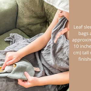 May include: A child holds a gray and brown fabric sleeping bag with bunny ears. Leaf sleeping bags are approximately 10 inches (25 cm) tall when finished.