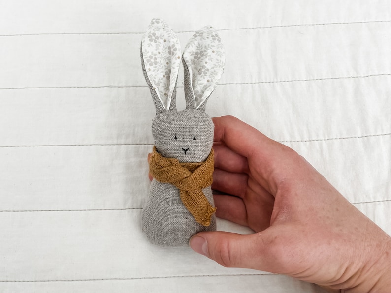 May include: A gray fabric bunny with a brown scarf. The bunny has small black eyes and a stitched nose. The bunny is held in a hand.