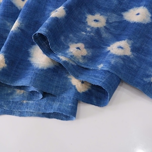 May include: Folded blue fabric with a white circular pattern. The fabric has a textured appearance, possibly linen or cotton. The design is similar to tie-dye or shibori techniques. The fabric is folded over on itself.