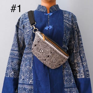 May include: A black and white patterned fanny pack with a gold zip and black leather trim. The bag is worn across the body. The person is wearing a blue patterned shirt with a mandarin collar. The number "#1" is in the upper left corner.