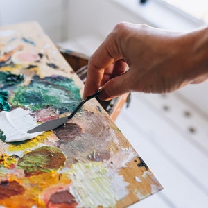 May include: A close-up of a wooden artist's palette with a variety of colorful oil paints. A hand is using a metal palette knife to mix the white paint.
