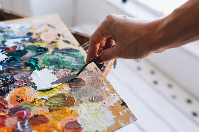 May include: A close-up of a wooden artist's palette with a variety of colorful oil paints. A hand is using a metal palette knife to mix the white paint.