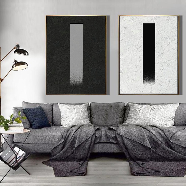 May include: Two abstract paintings with a vertical gray and black stripe on a black and white background. The paintings are framed in gold and hung on a white wall above a gray couch.
