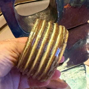May include: A cylindrical ceramic object featuring a ribbed, textured surface with alternating vertical bands of glossy brown and cream-coloured glaze. The piece has a smooth, reflective finish and a hollow centre, showcasing a layered, earthy colour palette.