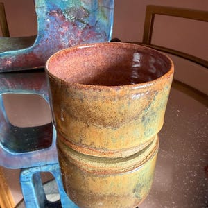May include: A cylindrical ceramic bowl with a glossy finish, featuring a gradient of warm amber, golden yellow, and earthy brown tones. The interior rim displays a soft pinkish-red hue, and the exterior surface shows a mottled, textured glaze.