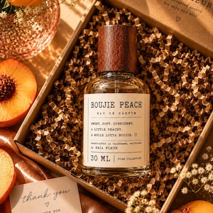 May include: A 30 ml glass perfume bottle with a dark wood cap, labeled Boujie Peach Eau de Parfum, nestled in a cardboard gift box filled with brown paper crinkle filler. The label includes the text Sweet. Soft. Confident. A little peachy.
