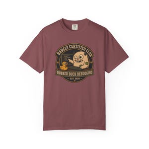 May include: A dusty rose short-sleeve t-shirt featuring a circular graphic with the text "BARELY CERTIFIED CLUB" and "RUBBER DUCK DEBUGGING EST. 2026". The design shows a beige monster at a computer with a yellow rubber duck wearing glasses.