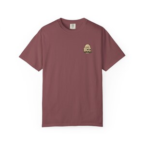 Puede incluir: Una camiseta de manga corta en color rosa viejo con un pequeño gráfico en el pecho izquierdo. El gráfico muestra un personaje de dibujos animados sobre las letras BCC en fuente negrita, con texto adicional debajo. Tiene cuello redondo.
