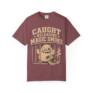 May include: A maroon short-sleeve t-shirt featuring a graphic of a beige, furry monster with mismatched eyes inside a cage. The text above reads CAUGHT RELEASING MAGIC SMOKE. The monster is standing next to a small workbench with tools and smoke.