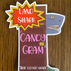 May include: A sticker with a cartoon shark peeking out from behind a door. The door has a sign that says "Land Shark "Candy Gram" That Clever Shark".