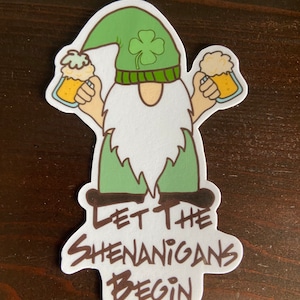 May include: A white sticker with a cartoon gnome holding two mugs of beer. The gnome is wearing a green hat with a shamrock on it. The text "Let the Shenanigans Begin" is written in brown below the gnome.