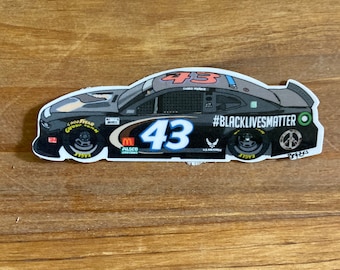 May include: A black and grey racing car with the number 43 on the side. The car has various sponsors including McDonald's, Alsco Uniforms, and Goodyear. The car also has the text "#BlackLivesMatter" on the side.
