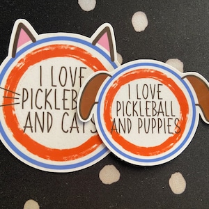 May include: Two stickers with the text "I LOVE PICKLEBALL AND CATS" and "I LOVE PICKLEBALL AND PUPPIES". The cat sticker has a cat head outline, and the dog sticker has a dog head outline. Both stickers have a red and blue circle design.