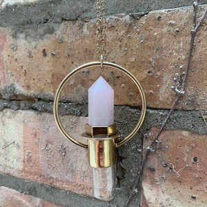 Könnte beinhalten: Eine goldfarbene Halskette mit einem durchsichtigen Glasfläschchen, einem rosa Kristall und einem kreisförmigen Anhänger. Der Kristall hat die Form eines spitzen Obelisken. Die Halskette hängt vor einer Backsteinmauer.