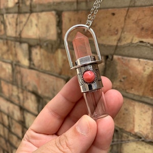 May include: A silver-toned necklace featuring a clear glass vial pendant with a pink crystal and a decorative silver band with a coral-colored stone. The pendant hangs from a delicate chain.