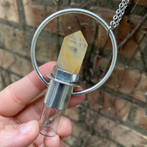 Könnte beinhalten: Eine silberfarbene Halskette mit einem durchsichtigen Glasfläschchen und einem gelben Kristallpunkt. Das Fläschchen wird in einem silbernen Ring gehalten und ist an einer Kette befestigt. Der Kristall ist hellgelb.