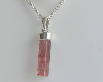 May include: A pendant necklace featuring a raw, cylindrical pink tourmaline crystal suspended from a silver-toned metal cap. The pendant hangs from a delicate, twisted silver-toned rope chain, showcasing the natural vertical striations of the stone.
