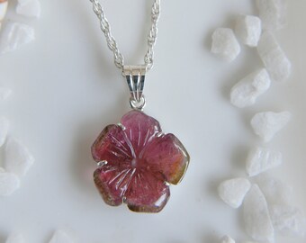 May include: A silver chain necklace featuring a carved pink tourmaline gemstone pendant shaped like a five-petaled flower. The pendant is held by a silver bail and displays natural color variations ranging from deep magenta to light olive green.