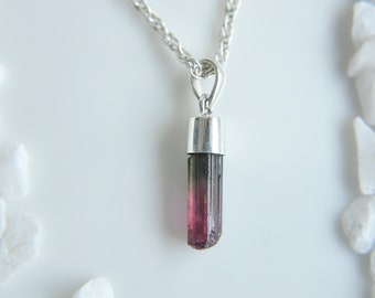 Puede incluir: Un collar con colgante que presenta un cristal de turmalina en bruto orientado verticalmente, con un degradado de verde oscuro en la parte superior que pasa a un rosa magenta vibrante en la base, sujeto por una tapa de metal plateado en una cadena.