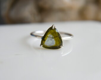 May include: A silver ring featuring a triangular, faceted gemstone with a gradient of olive green at the edges transitioning to a pale, translucent centre. The stone is held in place by three small metal prongs on a thin, smooth, polished silver band.